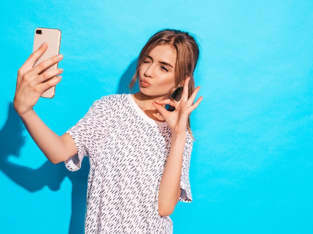 사진 selfie를 복용 쾌활 한 젊은 여자의 초상화입니다. 스마트 폰 카메라를 들고 아름 다운 여자입니다. 스튜디오에서 파란색 벽 근처 포즈 모델 미소. 확인 표시를 보여줍니다. 윙크하고 오리 얼굴을 만듭니다.