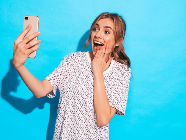 写真selfieを撮る陽気な若い女性の肖像画。スマートフォンのカメラを保持している美しい少女。スタジオの青い壁に近いポーズモデルの笑みを浮かべてください。びっくりしたモデル