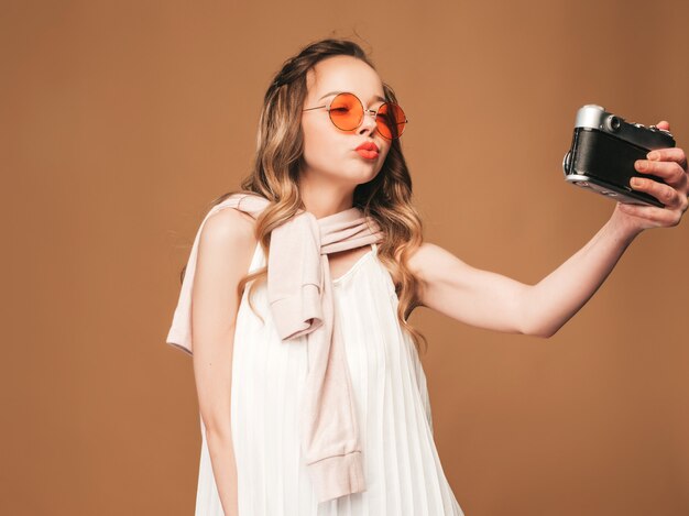 Portrait of cheerful young woman taking photo with inspiration and wearing white dress. Girl holding retro camera. Model posing.Making selfie