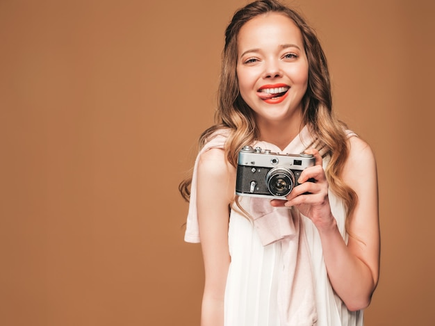 Portrait of cheerful young woman taking photo with inspiration and wearing white dress. Girl holding retro camera. Model posing