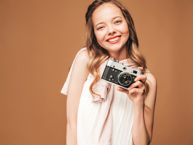 Portrait of cheerful young woman taking photo with inspiration and wearing white dress. Girl holding retro camera. Model posing