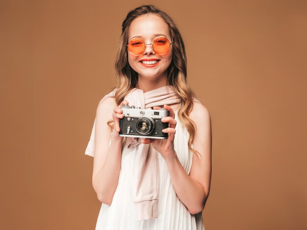 Portrait of cheerful young woman taking photo with inspiration and wearing white dress. Girl holding retro camera. Model posing