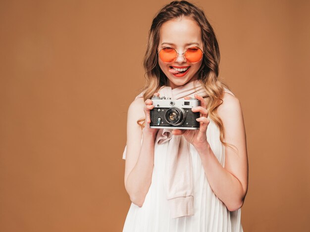 Portrait of cheerful young woman taking photo with inspiration and wearing white dress. Girl holding retro camera. Model posing