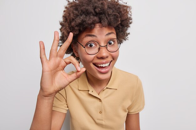 巻き毛の陽気な若い女性の肖像画は大丈夫ゼロ記号を示していますすべてがうまく見えることを保証します白い背景に対して丸い眼鏡カジュアルなTシャツのポーズを喜んで着ています。とても良い