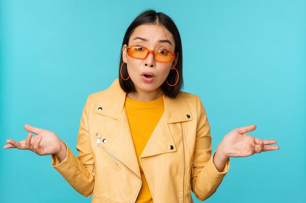 Portrait of chinese girl in sunglasses looks confused shrugs shoulders and staring puzzled at camera standing over blue background