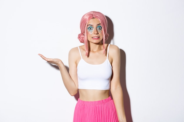 Портрет невежественной милой девушки в розовом парике, пожимающей плечами, ничего не подозревающей, стоящей в костюме на Хэллоуин с ярким макияжем.