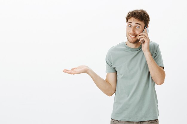 Portrait of clueless handsome Caucasian man with fair hair, shrugging with spread palm and talking on smartphone, being unaware and questioned