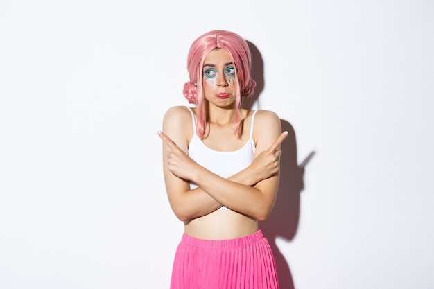 Портрет невежественной глупой молодой девушки в розовом парике и хеллоуинском костюме, нерешительно пожимая плечами и надувая губы, указывая пальцами в сторону, стоя на белом фоне