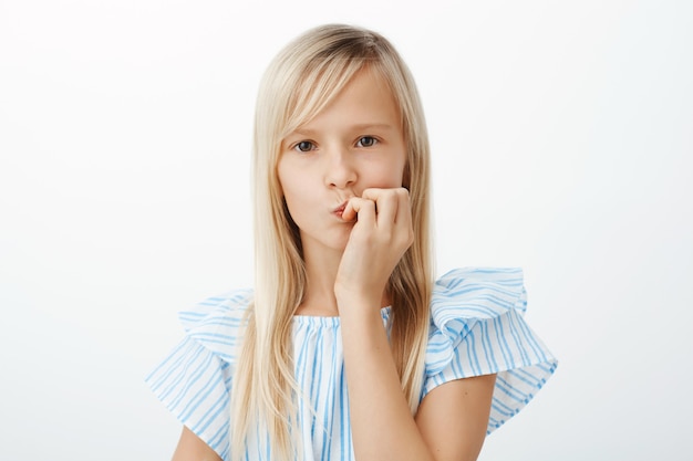 Portrait of concerned focused little girl with long blond hair, folding lips and biting fingers, staring, spacing out during scolding or thinking about personal problems over gray wall