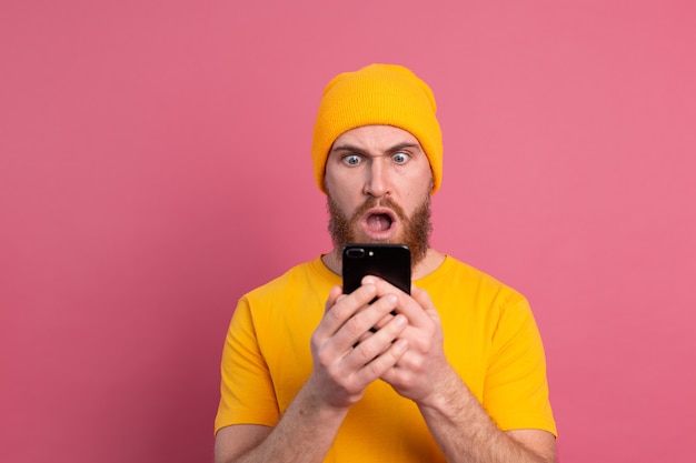 Portrait of concerned shocked mature bearded male gasping unhappy holding smartphone reading strange and disturbing message on pink