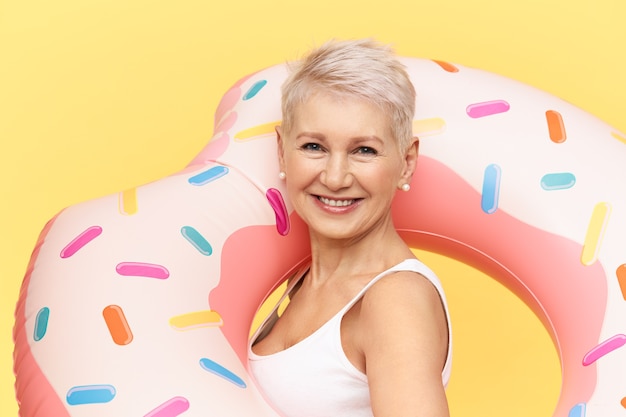 Portrait of confident attractive European female with pixie stylish haircut posing against yellow background, carrying pink inflatable swimming circle, spending hot sunny day by sea or pool, smiling
