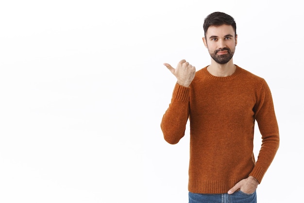 Portrait of confident bearded handsome man in sweatshirt pointing thumb left showing way with pleased satisfied expression recommend good store for real men white background