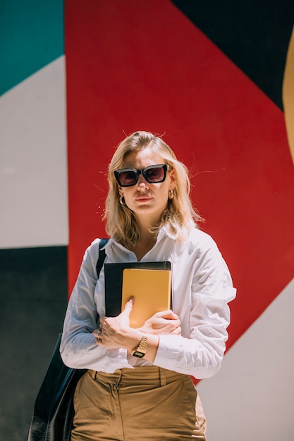 Free photo portrait of a confident blonde young businesswoman standing against colored painted wall