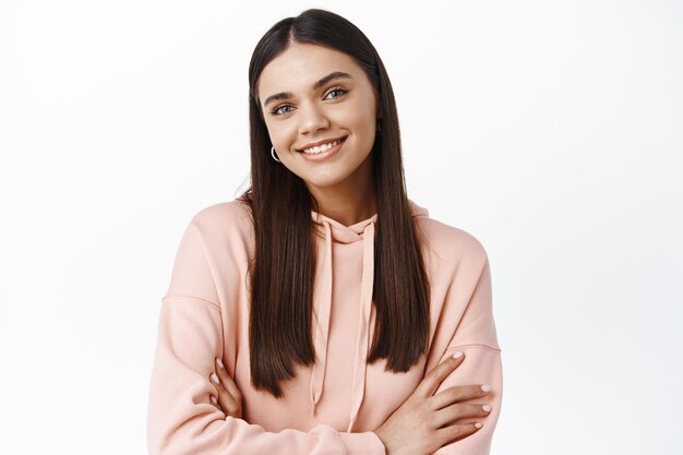 Portrait of confident brunette woman looking happy and self-assured, cross arms on chest and smile at front, standing against white wall