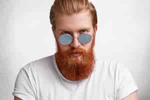 Free photo portrait of confident brutal male with stylish haircut, has thick long fuzzy red beard and mustache, wears round shades and white casual t shirt, poses alone against concrete wall.