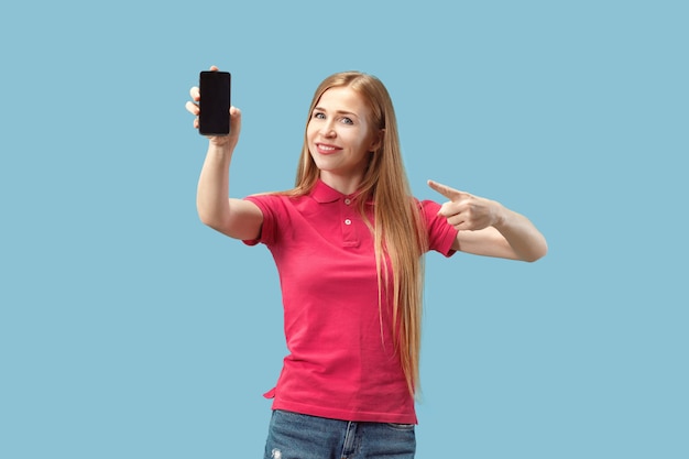 Portrait of a confident casual girl showing blank screen mobile phone isolated over blue background