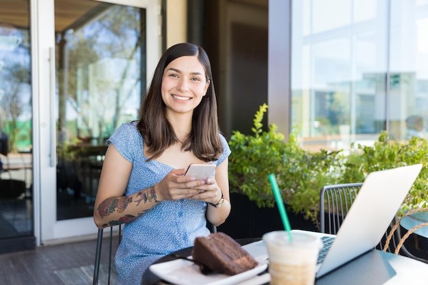 Portrait of confident female blogger using smartphone and laptop while having desert in cafe