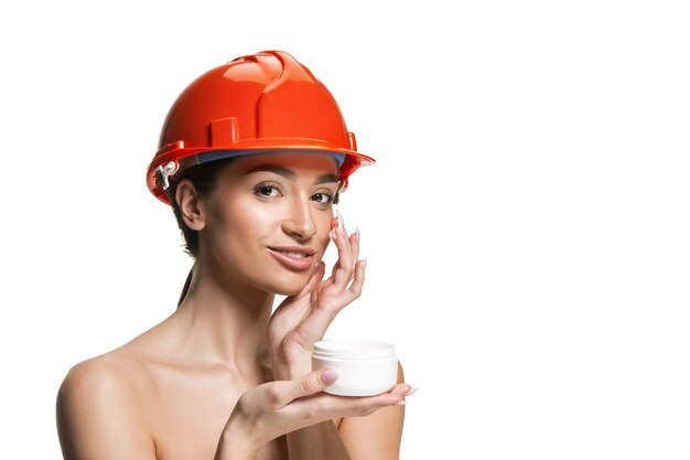 Портрет уверенно женского счастливого улыбающегося работника в оранжевом шлеме. Женщина изолированная на белой стене. Красота, косметика, уход за кожей, защита кожи и лица, косметология и концепция кремов