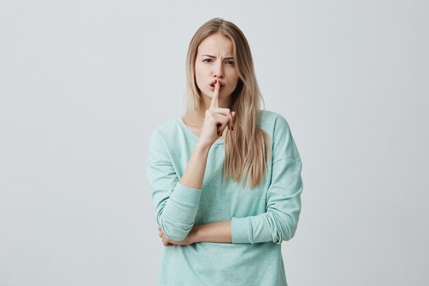 Portrait of confident female with long blonde hair wears blue clothes, shows silence sign, asks to keep private information confidential, has serious expression. Secret gesture