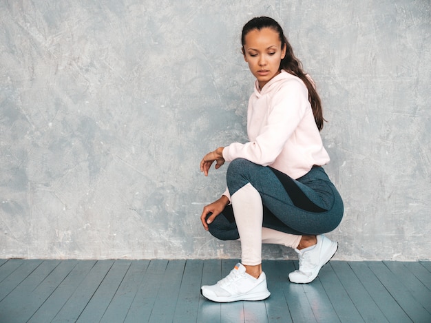 Free photo portrait of confident fitness woman in sports clothing looking confident.female sitting in studio near gray wall