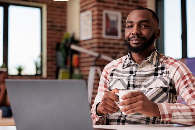Portrait of confident freelance programmer looking at camera taking a break from coding holding a cup of coffee. African american student relaxing at home watching social media content on laptop.