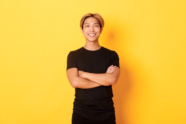 Portrait of confident handsome asian man smiling satisfied, standing over yellow wall in black clothes.