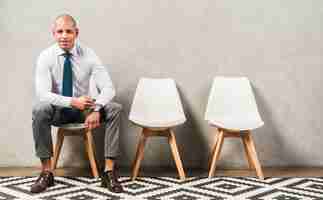 Free photo portrait of a confident happy young businessman sitting on chair in front of grey wall