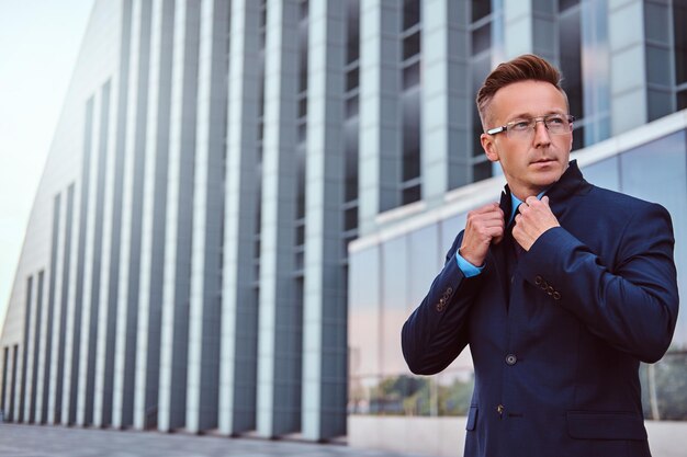 Portrait of a confident man in elegant suit and glasses correct his jacket while standing against a skyscraper background.