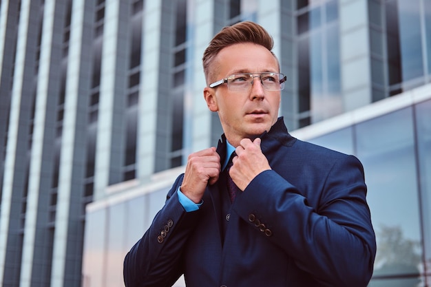 Free photo portrait of a confident man in elegant suit and glasses correct his jacket while standing against a skyscraper background.