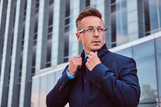 Portrait of a confident man in elegant suit and glasses correct his jacket while standing against a skyscraper background.