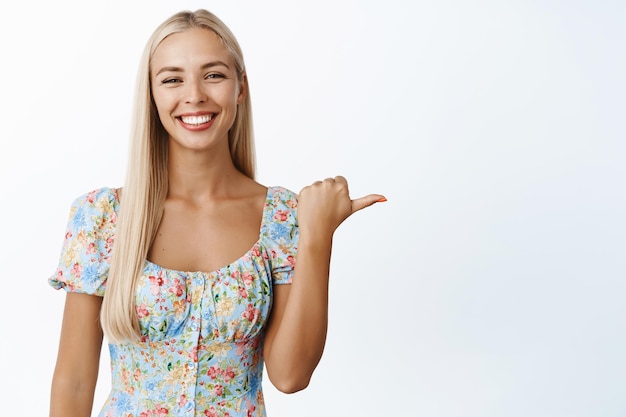 Foto gratuita ritratto di donna bionda sorridente sicura che mostra l'annuncio che punta a destra sull'annuncio di vendita del logo in piedi su sfondo bianco
