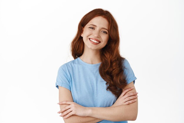 Portrait of confident smiling girl with red curly hair, cross arms on chest and looking determined, feeling relaxed like real professional, standing in t-shirt against white background.