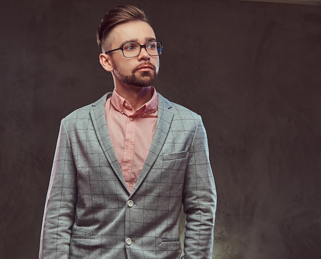 Портрет уверенного в себе стильного бородатого мужчины с прической и очками в сером костюме и розовой рубашке, позирующего в студии. Изолированные на сером фоне.