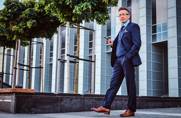 Foto gratuita ritratto di un uomo d'affari fiducioso ed elegante vestito con un abito elegante tiene uno smartphone e distoglie lo sguardo mentre si trova all'aperto sullo sfondo del grattacielo.