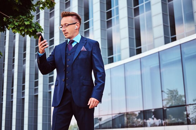 エレガントなスーツに身を包んだ自信を持ってスタイリッシュなビジネスマンの肖像画は、高層ビルの背景に屋外に立っている間、スマートフォンを保持し、目をそらします。