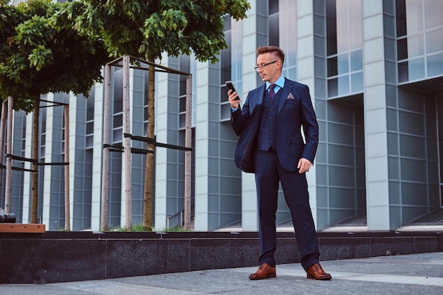 Portrait of a confident stylish businessman dressed in an elegant suit using a smartphone while standing outdoors against a skyscraper background.