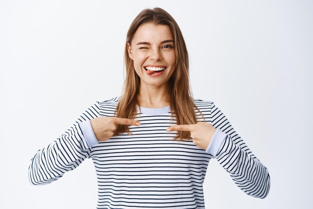 Portrait of confident woman pointing at herself and winking willing to be chosen or picked nominating her selfpromoting while standing over white background