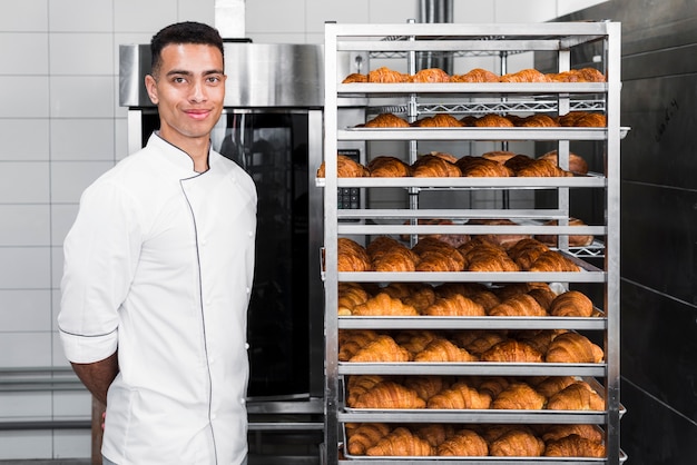 Portrait of a confident young male baker standing near the baked croissant shelves