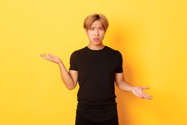 Портрет смущенного и раздраженного азиатского парня, раскинувшего руки в стороны, ничего не понимающего, стоящего у желтой стены