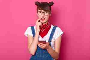 Free photo portrait of confused cute girl wears denim overalls, casual white t shirt, holds mobile phone in hand, keeps fore finger on lip