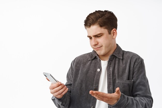 Portrait of confused guy stares at smartphone screen and shrugs, cant understand message, looking at mobile phone notification indecisive, standing on white