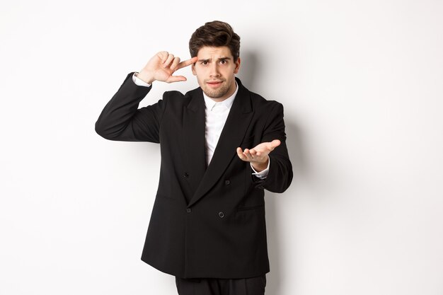 Portrait of confused handsome businessman scolding employee, pointing at head and camera, standing disappointed over white background