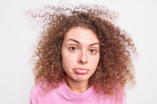Free photo portrait of confused indignant with curly hair purses lower lip looks surprisingly  feels embarrased as hears unexpected news dressed in casual jumper isolated over white wall