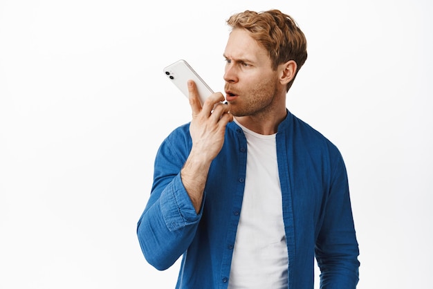 Portrait of confused redhead man hold phone close to lips shouting at speakerphone and frowning annoyed recording voice message with angry face standing over white background