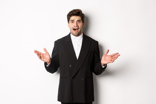 Portrait of confused and worried handsome man in suit, looking at something strange, spread hands sideways and standing puzzled against white background
