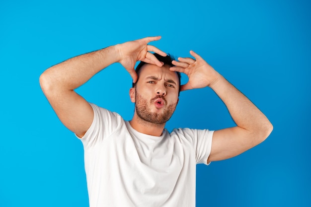 Portrait of a confused young man holding hands on his head over blue background