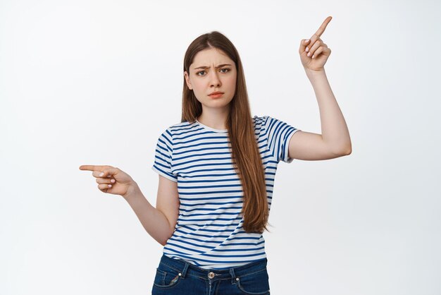 Portrait of confused young woman pointing up and left with indecisive frowning face puzzled what to choose standing over white background