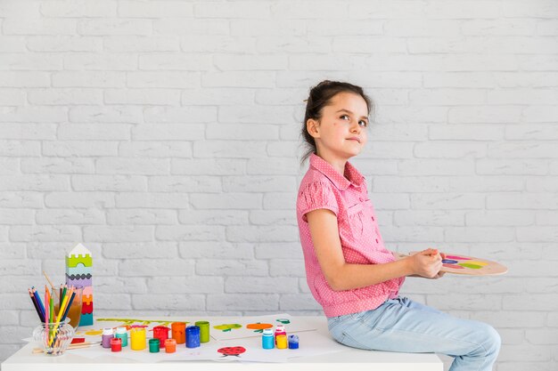 Portrait of a contemplated girl sitting on the white table mixing the watercolor on palette