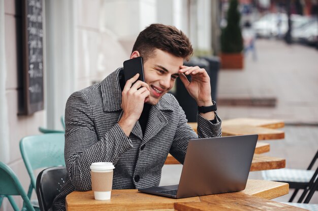 Portrait of content guy drinking takeaway coffee in street cafe, working with notebook and having pleasant mobile conversation