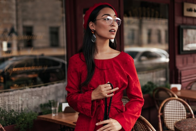 Il ritratto di una donna abbronzata fresca in berretto rosso, abito elegante e occhiali tiene una borsa nera e posa all'esterno